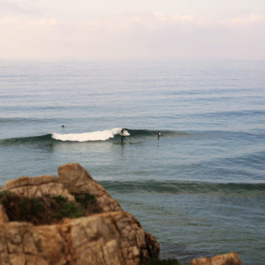 ein Surfer auf einer Welle aus Vogelperspektive und Fels im Vordergrund