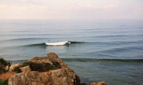 ein Surfer auf einer Welle aus Vogelperspektive und Fels im Vordergrund