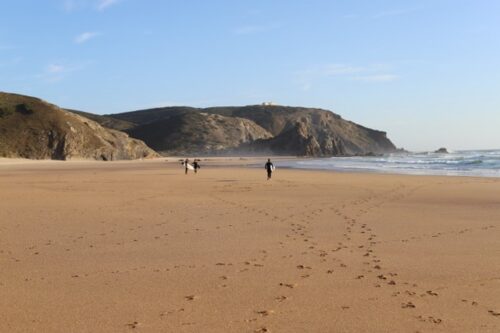 Surfer laufen in der Ferne an einem Strand entlang. Klippen im Hintergrund.